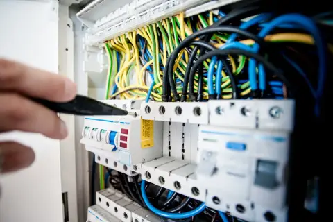 Ręka z narzędziem przy bezpiecznikach. W tle plątanina kolorowych przewodów, grożąca zwarciem instalacji elektrycznej.